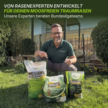 Ein lächelnder Mann mit Brille und grünem Terrauno-Hemd kniet im Gras neben drei Moosfrei Soforthilfe Set-Taschen von Terrauno, dahinter ein Garten und eine Holzkonstruktion. Der Text hebt die von Experten entwickelte Moosbekämpfung für moosfreie Rasenflächen hervor.