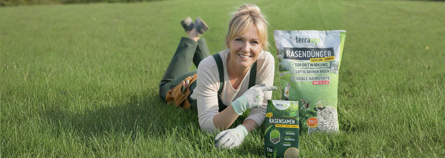 Eine lächelnde Frau in Gartenarbeiterkleidung liegt auf grünem Gras und zeigt auf Säcke mit Rasendünger und Grassamen. Die Produkte sind vor ihr aufgereiht, und sie trägt Handschuhe und hat Gartengeräte dabei.