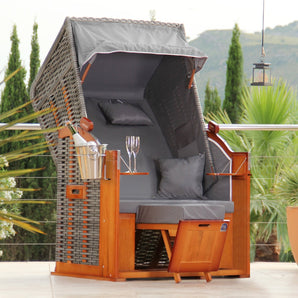 Der DERKORB Der Klassiker 1-Sitzer ist ein stilvoller Strandkorb aus Kiefernholz und Polyrattan mit Baldachin, grauen Kissen, Fußstützen und Holztabletts für Getränke - ideal zum Entspannen im Freien inmitten von üppigem Grün.