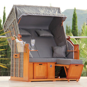 Der DERKORB Der Klassiker 2-Sitzer ist ein moderner Strandkorb aus Polyrattan und Kiefernholz mit einem Baldachin, grauen Kissen, eingebauten Fußstützen, Beistelltischen mit Gläsern und einem Eiskübelhalter - perfekt für Ihre Terrasse.