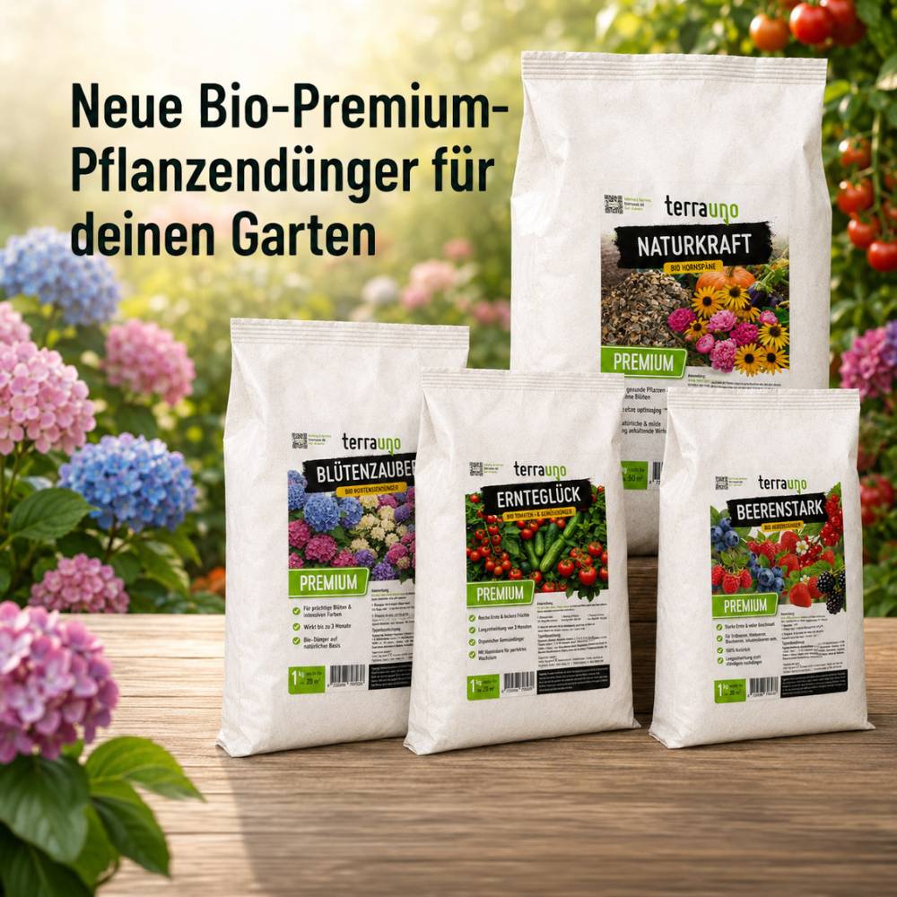 Auf einem Holztisch im Freien stehen vier Säcke mit Bio-Premium-Pflanzendünger, umgeben von bunten Blumen. Auf einem grünen Etikett steht „NEU“ und in Fettdruck: „Neue Bio-Premium-Pflanzendünger für deinen Garten.“.