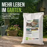 Ein Beutel „TerraUno Premium Wildvogelfutter“ steht auf einem Holztisch im Freien, daneben eine grüne Gießkanne und im Hintergrund blühende Blumen. Der Text hebt dieses hochwertige, schalenlose Ganzjahresfutter als das ideale Vogelfutter für den Garten hervor, mit dem sich Gartenvögel das ganze Jahr über füttern lassen.